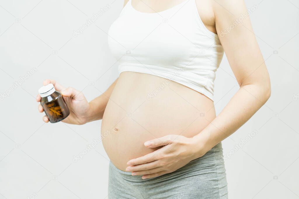 Selective focus of pregnant woman with Medicine bottle in Hand Touching Belly Standing Over Gray  Background,Healthy millennial women taking antioxidant medicine vitamins, healthcare concept.