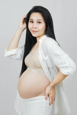 Genç, mutlu, gülümseyen Asyalı kadının portresi, hamilelik sağlık sigortası bebek için hazırlanıyor. Genç anne arasında annelik, sarılan göbek, gri arka planda izole edilmiş.