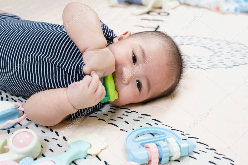 Asian baby four or five months old having teeth growing issues teething pain while holding a bite toy looking to the camera lying on the bed with teether at home