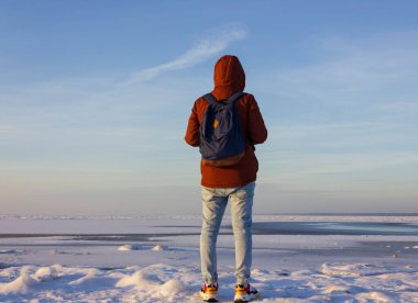 Yolculuk sırasında sırt çantalı erkek turistin donmuş körfezin kıyısında duruşu. Sırt çantalı kırmızı ceketli bir gezgin doğayı keşfeder. Gezgin yaşam tarzı..