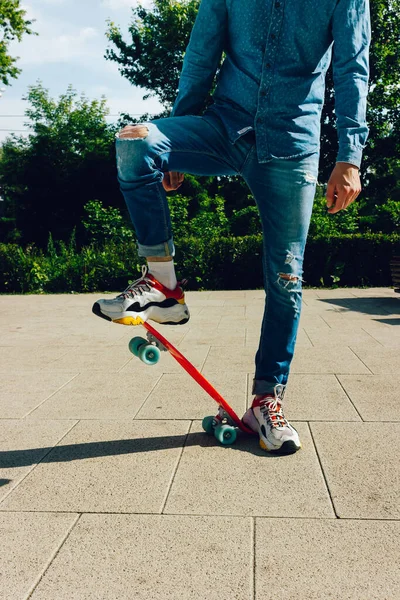 Mladík v roztrhaných džínách stojí s penny board v parku. letní aktivity skateboarding — Stock fotografie