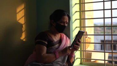 Yüzü koruyucu maskeli Hintli kadın izolasyon sırasında tabletten haber okuyor. 
