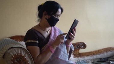 Yüzü koruyucu maskeli Hintli kadın izolasyon sırasında tabletten haber okuyor. 