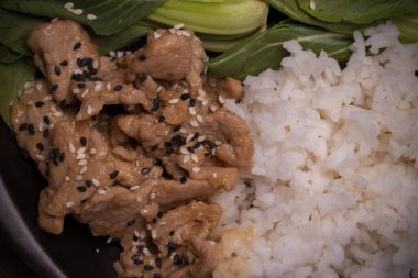 Teriyaki domuz eti üzerine susam ve sebze serpilmiş ve siyah tabakta pilav..