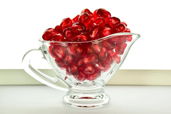 Dans un bol en verre transparent sont versés juteux rouge vif graines de fruits de grenade. Sur fond blanc — Photo