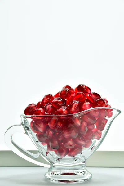 Dans un bol en verre transparent sont versés juteux rouge vif graines de fruits de grenade. Sur fond blanc — Photo
