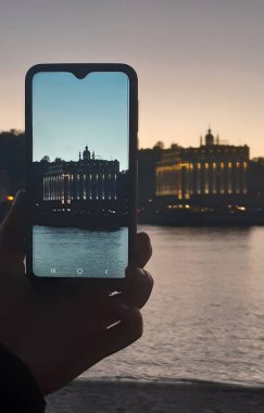 Bir el, nehrin karşısındaki binanın fotoğrafını cep telefonuyla çekiyor. Bina telefonda temiz, arka planda da da bulanık..