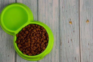 Gri ahşap tahtalardan oluşan arka planda, kedi ve köpekleri beslemek için kuru yiyeceklerle dolu yeşil bir plastik tabak yatıyor. Yazı için sağda bir yer var.