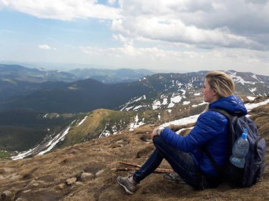 Sırt çantalı sarışın bir kız, bir dağın zirvesinde oturuyor, gökyüzünün arka planında ve karla kaplı dağlarda.