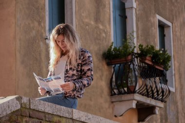 Sarışın bir kız, çiçeklerle dolu eski bir balkonun arka planındaki taş korkulukta oturuyor ve dergi okuyor.