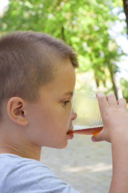 Little boy drinks from a large plastic clear glass of lemonade close up. Shot from the side. The background is blurred