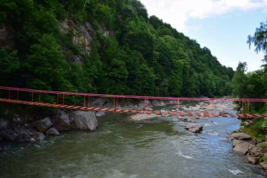 Dağlık yeşil bitki örtüsünde bir dağın arka planına karşı dağ nehri üzerindeki kayalık kıyılar arasında insanlar olmadan asılmış halat köprü.
