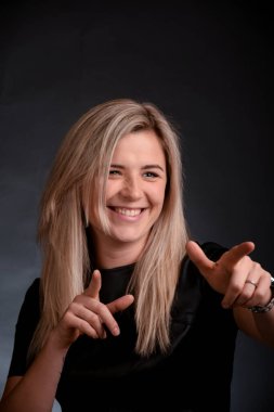 Attractive blonde girl laughs and shows her fingers to the side on a dark background. Photographed in the studio.