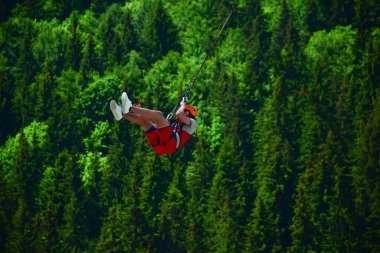 Miğferli genç bir adam bungee jumping 'den atladı ve şimdi bir ipe tutunuyor, yeşil bir ormanın bulanık arka planına karşı kendini bir spor kamerasında sallıyor ve filme çekiyor.