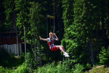 Miğferli genç bir adam bungee jumping 'den atladı ve şimdi bir ipe tutunuyor, yeşil bir ormanın bulanık arka planına karşı kendini bir spor kamerasında sallıyor ve filme çekiyor.