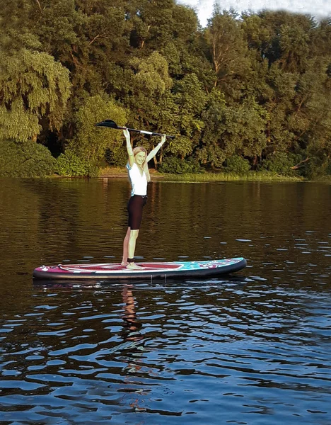 一个漂亮的姑娘站在一块游泳板上 在河水 森林和灿烂的阳光的映衬下 高高地高举着桨 — 图库照片