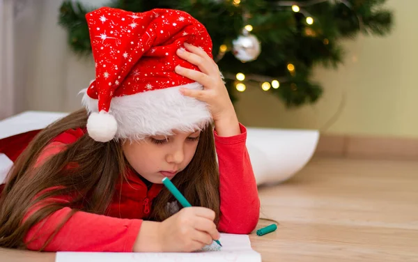 Bir kızın portresi. Kırmızı Noel Baba şapkalı, Noel Baba 'ya evde Noel ağacının yanında mektup yazan tatlı küçük bir kız. Yeni yıl 2021..