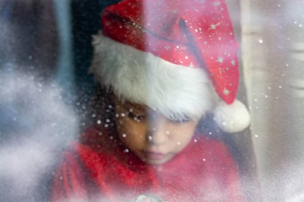 Sarı saçlı, kırmızı Noel pijamalı ve Noel Baba şapkalı küçük beyaz kız. Büyük pencerede duruyor ve karlara bakıyor. Noel arkaplanı.