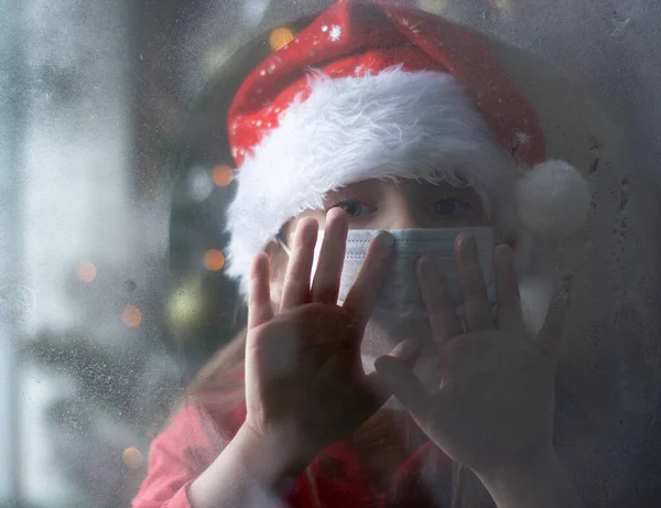 Una niña europea con máscara médica, sombrero y traje de Santa mira a través de una ventana cubierta de nieve. Árbol de Navidad en el fondo. Cara en desenfoque. Copiar espacio. — Foto de Stock