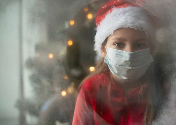 Tıbbi maskeli, şapkalı ve Noel Baba kostümlü küçük bir Avrupalı kız karla kaplı pencereden bakıyor. Arka planda Noel ağacı var. Yüzü dağılmış. Boşluğu kopyala.