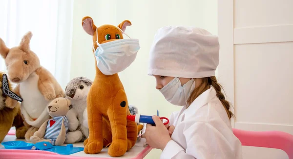 Beyaz bir sağlık önlüğü ve tıbbi maske takmış küçük bir kız, bir doktoru oynuyor ve çocuk odasının arka planında çeşitli hastalıklara karşı çeşitli aşılar yapıyor..