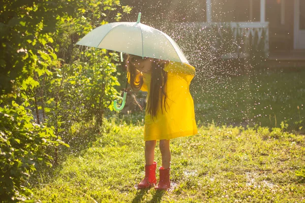A happy girl with a green umbrella under the summer rain. The girl is dressed in a yellow raincoat and enjoys the rain. The child plays in nature in the fresh air. — Stock Photo, Image