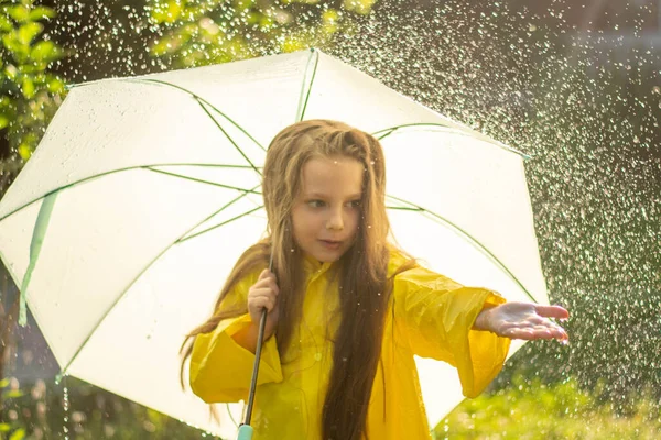 A happy girl with a green umbrella under the summer rain. The girl is dressed in a yellow raincoat and enjoys the rain. The child plays in nature in the fresh air.