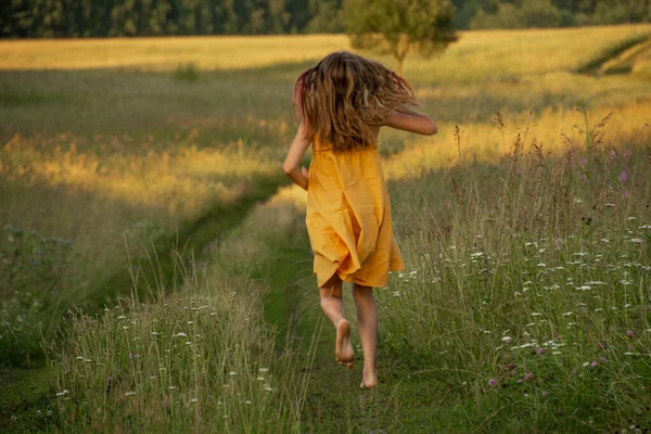 A girl in a yellow dress runs away on the road to a field at sunset. — Stock Photo, Image