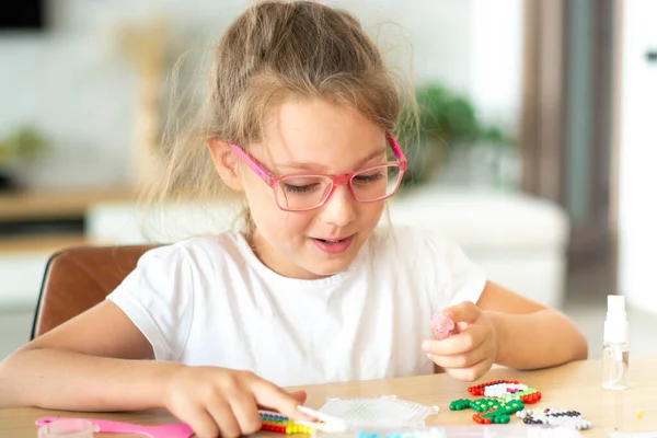 Una chica sentada en una mesa hace figuras de cuentas de colores. Aqua juego de mosaico. El concepto de ocio infantil. Imágenes de stock libres de derechos