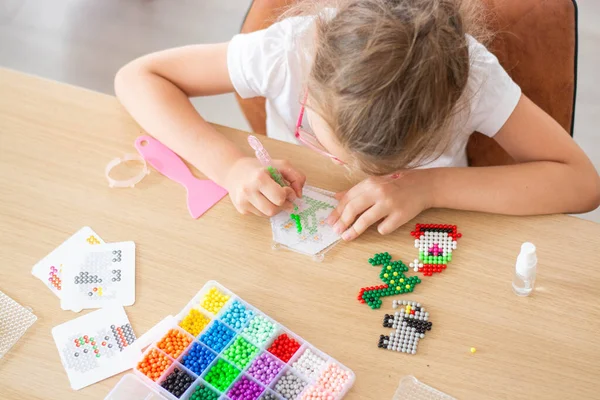 Una chica sentada en una mesa hace figuras de cuentas de colores. Aqua juego de mosaico. El concepto de ocio infantil. Tomado de arriba. Imagen de archivo