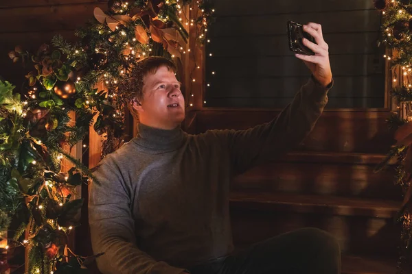 Caucasian man sits on wooden stairs and talks with his friend using app on smart phone. Stairs decorated with floral garland and Christmas lights for Christmas and New Year. Happy Holidays Theme.
