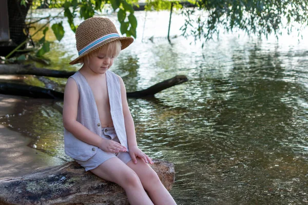 Sad and lonely little caucasian blond boy wearing straw hat sits by river. Fear of water. Mental Health Theme.