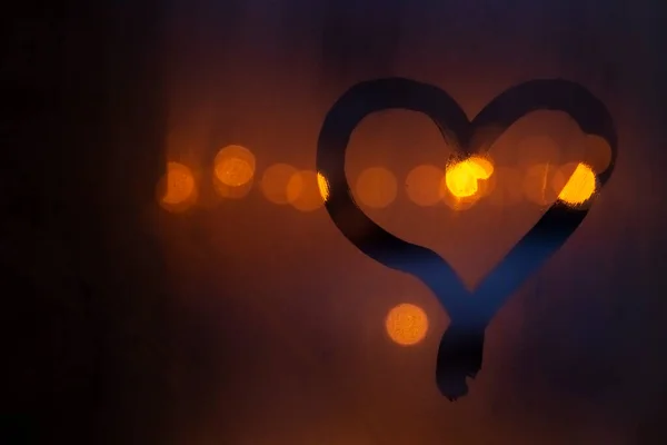 Close-up view of heart symbol drawing by finger on glass misted window. Orange blurred night background. Bokeh light. Selective focus. No people. Copy space for your text. Love and romance theme.