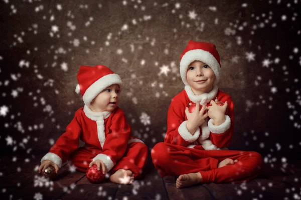 サンタクロースの衣装を着た2人の小さな子供がKoichnevの背景に座っていて 手にクリスマスツリーボールを持っています 白い雪片があちこちに降っている — ストック写真