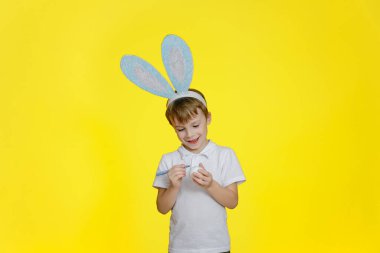 Sevimli bir çocuk sarı arka planda duruyor, kafasında ev yapımı tavşan kulakları, keçeli kalemle bir Paskalya yumurtası süslüyor.