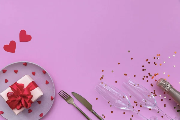 İki bardaklı şampanya ve sarı konfeti, pembe arka planda kalpleri olan kırmızı kurdeleli hediye paketi. Sevgililer Günü konsepti. Düz konum, üst görünüm, kopyalama alanı.