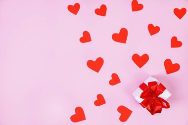 Kırmızı kurdeleli hediye kutusu, kırmızı kağıt kalplerin etrafında. Pembe arka plan. Sevgililer Günü konsepti, doğum günü. Düz konum, üst görünüm, kopyalama alanı.