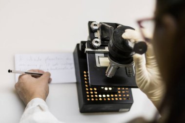 Bir bilimsel araştırmacı, mikroskop analizi yaparken ve bir kağıt parçasına notlar alırken,.