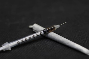 Kokain dolu bir şırınganın ön ve yakın görüntüsü, her ikisi de siyah bir yüzeyin aksine kenevir ve kokain kamışının üzerinde dinleniyor.