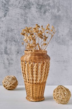 Kurumuş çiçekle örülmüş dallardan yapılmış kahverengi örülmüş vazo ve gri çimento arka planında beyaz ahşap masa üzerinde rattan topları. El yapımı, konfor ve ekolojik dekor kavramı. İç mimari.
