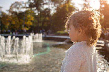 Küçük tatlı kız güneşli bir günde parktaki fıskiyelere bakıyor.