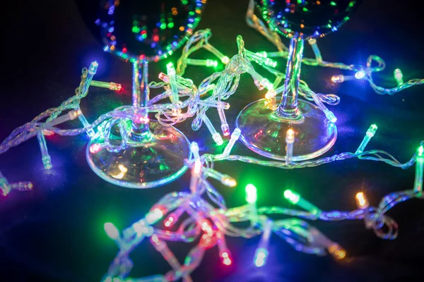 Χριστουγεννιάτικη λαμπερή πολύχρωμη γιρλάντα που αντανακλάται σε γυάλινα γυαλιά σε σκούρο φόντο. Πρωτοχρονιά εορτασμού. — Φωτογραφία Αρχείου