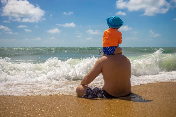 Dzień ojców. Tata z dzieckiem na ramionach siedzi na piasku i patrzy na morze. — Zdjęcie stockowe