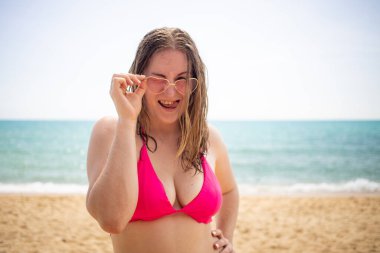 Gülümseyen, flört eden ve kameraya bakan pembe bikinili ve kalpli gözlüklü genç sarışın kız. Deniz plajı arka planda.