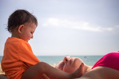 Küçük tatlı çocuk kumsalda, kumların üzerinde yatan annesinin yanında oturuyor. Arka planda güneşli bir günde ılık deniz.