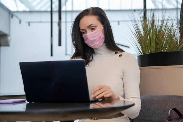 Mujer joven con mascarilla protectora funciona en el ordenador portátil en la cafetería... — Foto de Stock