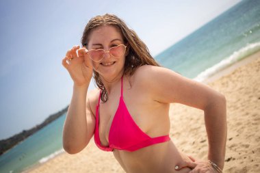 Sarı kız gülümsüyor ve neşeyle kameraya bakıyor, pembe bikini ve gözlük takıyor.