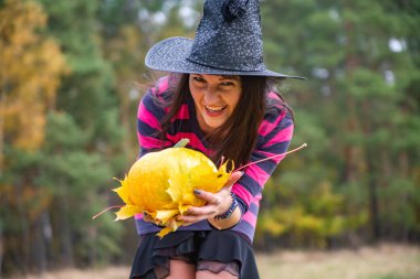 Cadı kadın gülerek balkabağını tutuyor ve sonbahar ormanında gülümsüyor. Cadılar Bayramı konseptini kapat.
