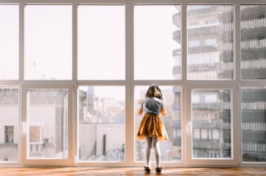 Bir kız pencerenin kenarında duruyor ve şehre bakan büyük bir panoramik pencereden dışarı bakıyor.