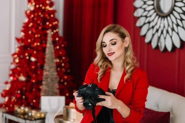beautiful woman photographer in red with a camera in her hands against the background of lights bokeh of a Christmas tree. Happy New Year 2021. Soft selective focus, art noise.